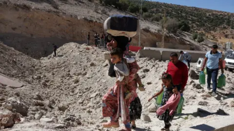 Reuters Sebuah keluarga dengan dua anak berjalan kaki - sang ibu membawa anak bungsunya dan beberapa tas - melintasi jalan berlubang di persimpangan Masna di Suriah pada 4 Oktober